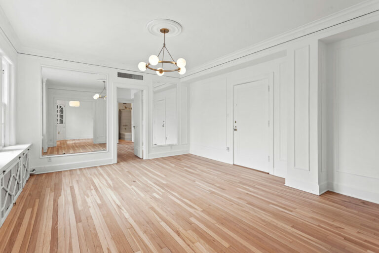 An empty room with laminated flooring, a chandelier, a huge mirror on one side of the room