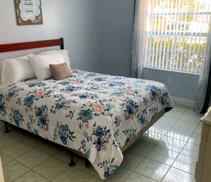 A double-sized mattress in a bedroom with powder blue painted wall, a window with blinds. and a board mounted against the wall