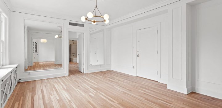 An empty room with laminated flooring, a chandelier, a huge mirror on one side of the room