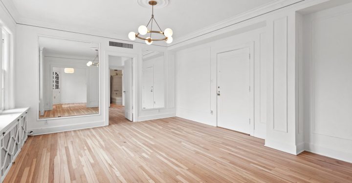 An empty room with laminated flooring, a chandelier, a huge mirror on one side of the room