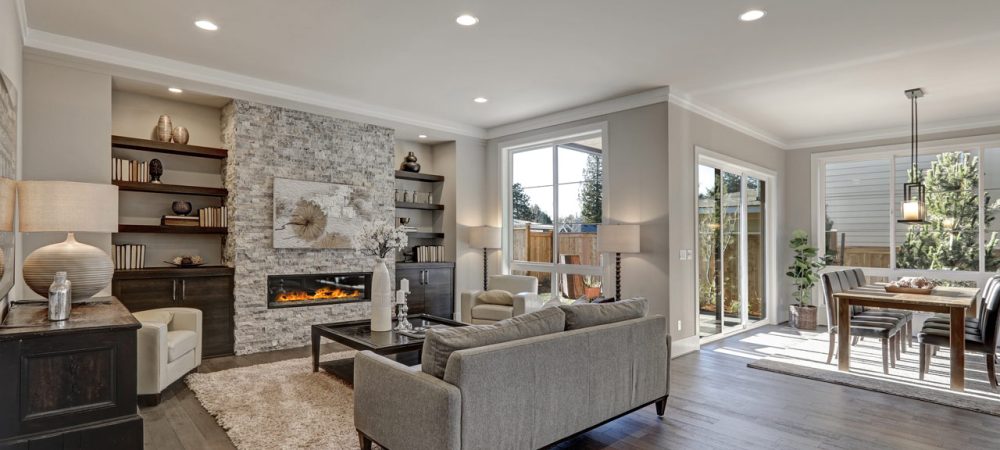 A seamless transition of a living room to a dining area: living room with gray couch, a fireplace, bookshelves, and several lamps. A dining area with a six-seater set and a hanging pendant light