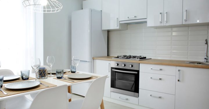 Modern kitchen interior with white hanging cabinets built around a refrigerator, a stove range, and an oven. A dining set with goblets, glasses, utensils, and plates.