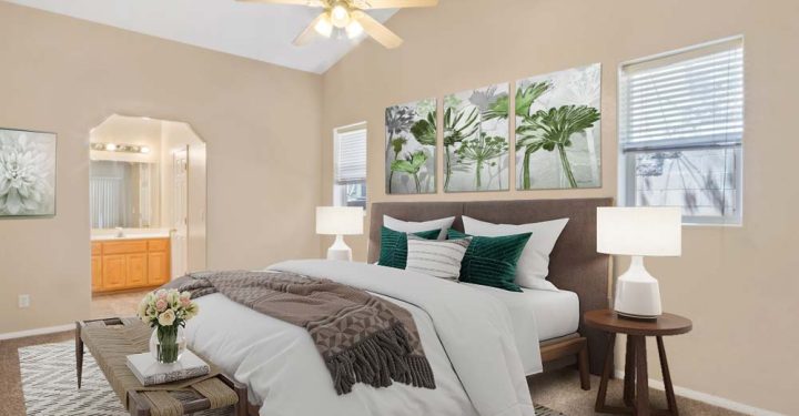 A bedroom with queen-size bed, side tables with lamps, a chandelier fan, windows with blinds, a wall art above the headboard, and a sneak peek to the bathroom