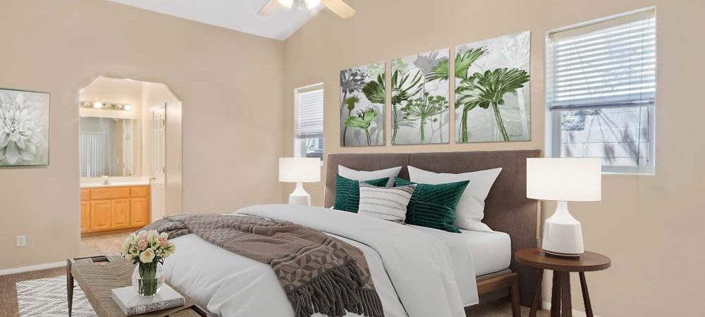 A bedroom with queen-size bed, side tables with lamps, a chandelier fan, windows with blinds, a wall art above the headboard, and a sneak peek to the bathroom