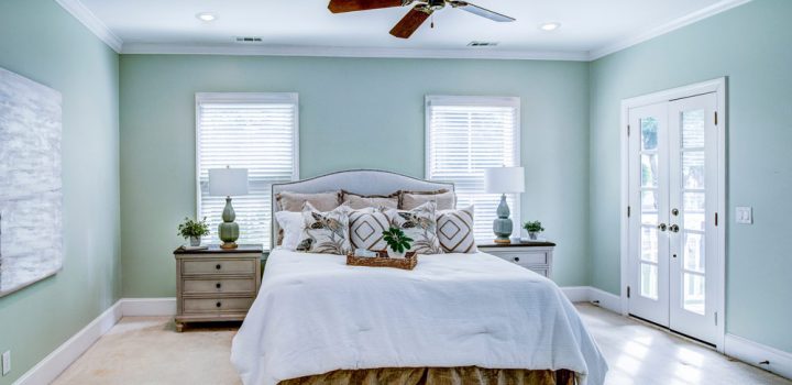 A queen-size bed, with two lamps and plant pots on top of wooden table for each side of the bed, windows with blinds behind the headboard, a ceiling fan, and a wall painted in cool mint green.