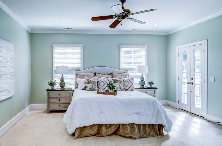 A queen-size bed, with two lamps and plant pots on top of wooden table for each side of the bed, windows with blinds behind the headboard, a ceiling fan, and a wall painted in cool mint green.