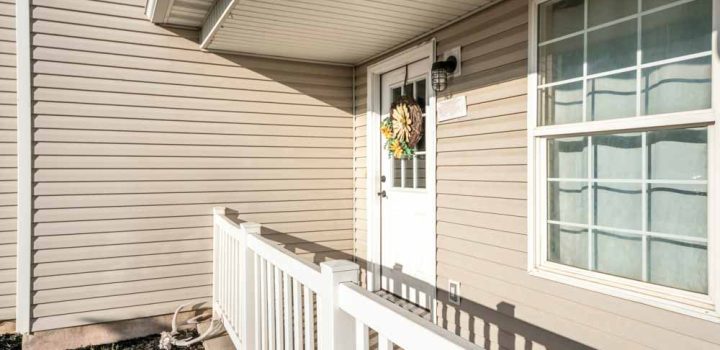 Front door exterior of a house with vinyl wall sidings. There is a white wooden railings at the front of a door with glass panel and wreath beside the double hung window.