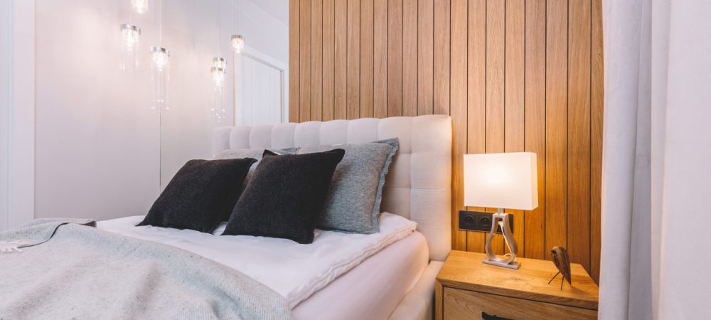 A double-sized white bed against wooden planks, and a bedside table with lamp